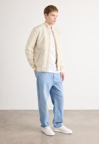 Beige bomber dzseki magas gallérral, alatta fehér póló, világoskék, laza szabású farmernadrág és fehér sportcipő. Simább anyag textúra.