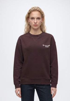 Femme blonde de face portant des boucles d'oreilles en or, un sweat marron foncé avec une inscription blanche sur la poitrine et un jean en denim foncé.