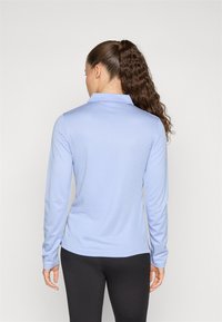 Camisa de manga longa azul clara com colarinho, tecido texturizado e fendas laterais. Corte slim, vista de trás, combinada com leggings pretas.