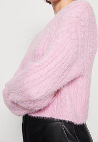 Suéter rosa esponjoso con un diseño de trenzas y mangas voluminosas. La textura parece suave y acogedora, contrastando con un atuendo negro.