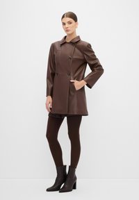 Bruine nepleder jas met een kraaglijn, dubbelknopen, zijzakken en driekwartmouwen, gecombineerd met donkere panty’s en enkellaarsjes.