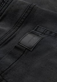 Tejido de mezclilla negro con una textura suave, que presenta un parche de cuero negro elevado en la cintura con el nombre de la marca y el año de establecimiento en relieve.