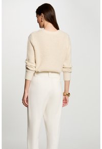 Beige gestrickter Pullover mit einem strukturierten Muster, rundem Halsausschnitt und gerippten Bündchen, kombiniert mit hochgelegten cremefarbenen Hosen.
