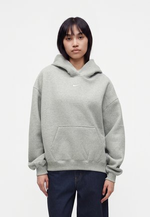 Femeie purtând un hanorac larg Nike gri deschis cu buzunar frontal și blugi bleumarin, stând în fața unui fundal alb simplu.