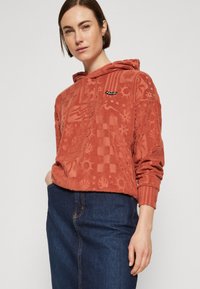 Sudadera de color óxido con patrones texturizados, que presenta un bolsillo tipo canguro y puños acanalados. Combinada con una falda de mezclilla.