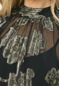 Top nero con tessuto trasparente decorato da motivi floreali metallici, colletto alto, dettagli a volant e un aspetto strutturato e stratificato.