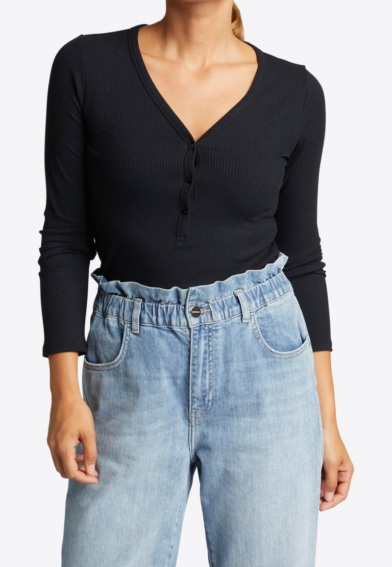 Schwarzes, geripptes Langarmshirt mit V-Ausschnitt und Knopfschluss, kombiniert mit hellblauen Denim-Jeans mit einem voluminösen Bund und Taschen.