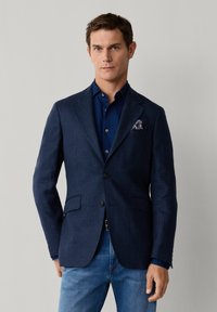 Mann trägt marineblauen karierten Blazer, dunkelblaues Hemd, blaue Jeans und gemustertes Einstecktuch vor einfachem hellem Hintergrund.