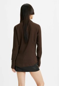 Chemise marron à motifs avec manches longues, ourlet droit et poignets à boutons, associée à une mini-jupe en cuir noir. Texture lisse.