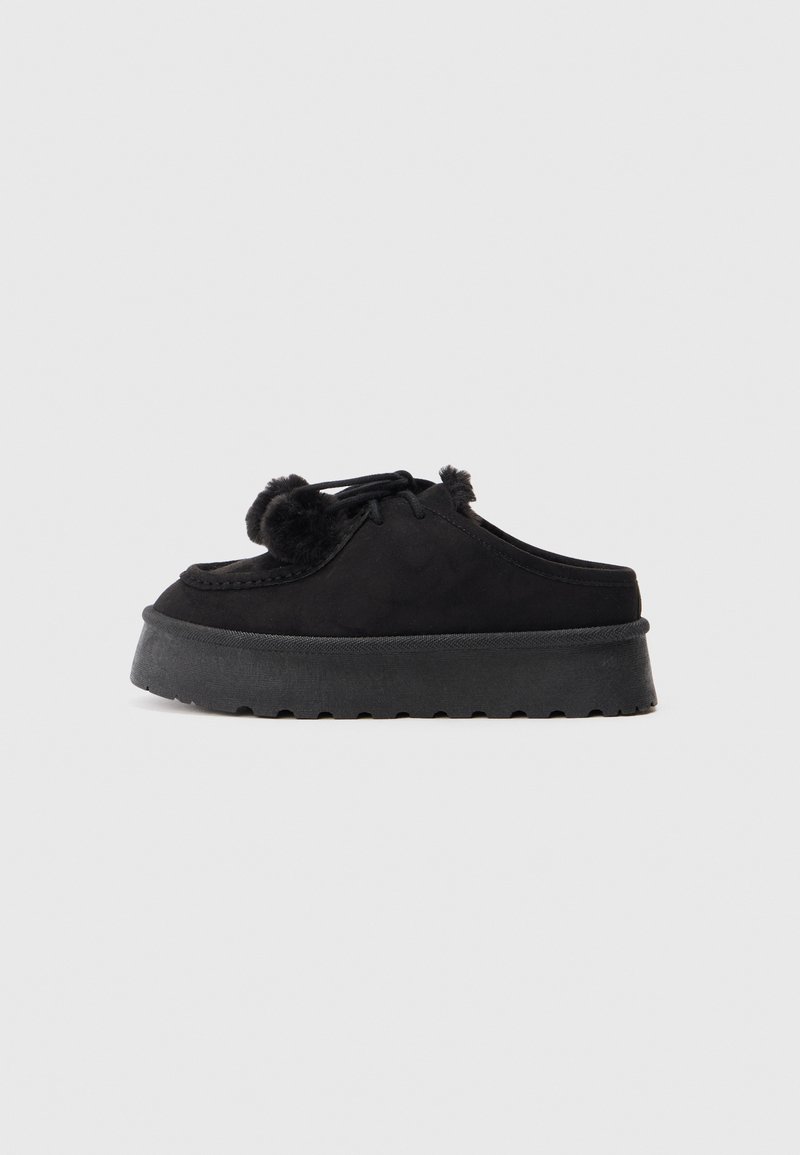 Des chaussures noires en daim à enfiler avec une semelle en caoutchouc épaisse, présentant un bout arrondi, des pompons décoratifs et des détails cousus.