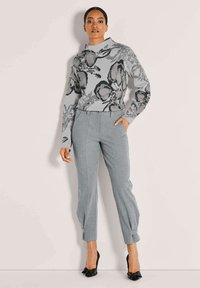 Vrouw in een grijze bloementrui, op maat gemaakte grijze broek met enkelstrikken en zwarte, puntige hakken, staande tegen een effen muur.