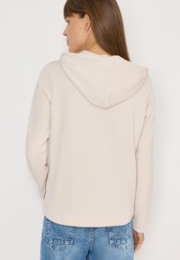 Jasnoróżowy sweatshirt z kapturem o miękkiej fakturze, z długimi rękawami i luźnym krojem. Widok z tyłu pokazuje regulowany kaptur oraz detale szwów.
