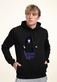 Sudadera negra con capucha que presenta un gráfico de un personaje con un traje a rayas y un águila, etiquetado como "Sombras Nocturnas y Cuervos." Tejido suave, bolsillo tipo canguro.