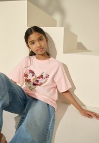 Rosa T-Shirt mit kurzen Ärmeln, das ein mehrfarbiges Pailletten-Herzdesign zeigt. Blaue Jeans im lässigen Schnitt. Der Hintergrund enthält hellfarbige Treppen.