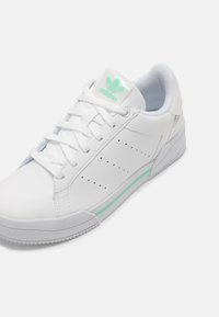 Bílé tenisky s hladkým syntetickým svrškem, zdobené akcenty v mátově zelené barvě, perforacemi a bílými tkaničkami. Gumová podešev s texturovaným gripem.