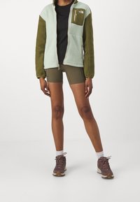 Ljusgrön fleecejacka med olivgröna ärmar och bröstficka, svart skjorta, olivfärgade shorts, bruna vandringsskor med krämfärgade sålar.