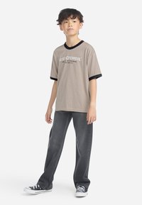 Beige t-skjorte med svart krage og mansjetter, prydet med hvit tekst. Kombinert med mørkegrå jeans og svarte joggesko med hvite såler.