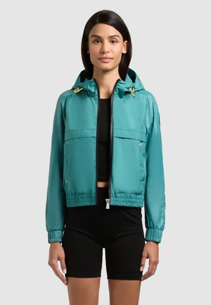 Femeie purtând o jachetă cu glugă turcoaz cu fermoar peste un top crop negru și pantaloni scurți strâmți negri, stând pe un fundal simplu, deschis la culoare.