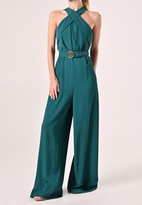 Mujer vestida con un mono sin mangas de color teal, con escote halter cruzado, piernas anchas y una cintura ceñida con un cinturón decorativo de hebilla dorada.