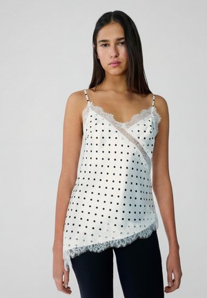 Mlada ženska z dolgimi temnimi lasmi, oblečena v belo svileno spodnjo majico s črnimi pikami in čipkastimi robovi, kombinirano s temnimi hlačami, stoji pred sivo podlago.