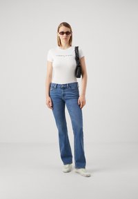 T-shirt bianco aderente con logo "TOMMY JEANS", jeans svasati in denim blu, borsa nera e occhiali da sole scuri, su uno sfondo neutro.