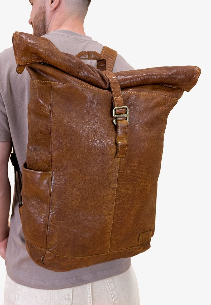 Sac à dos en cuir marron avec un dessus roulé, une sangle réglable, deux poches latérales et une finition douce et texturée. Design fonctionnel et durable.