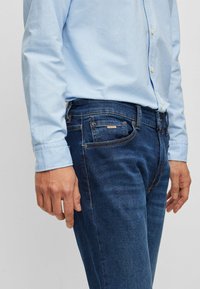 Denim jeans in dark blue with a slim fit, featuring subtle stitching, five pockets, and a branded tag on the front pocket. Styled with a light blue shirt.