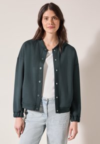 Tmavě zelená bomber bunda z hladké látky. Obsahuje patentky, žebrované manžety a klasický kulatý výstřih. Nositelná přes bílé tričko.