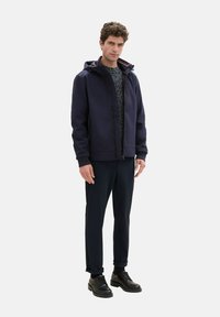 Navy Kapuzenjacke mit einem Reißverschluss vorne, gerippten Bündchen und Kordelzug. Kombiniert mit dunklen Hosen und schwarzen Schnürschuhen.