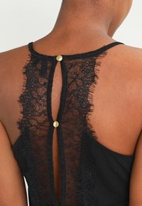 Robe noire avec un dos en dentelle et une ouverture en forme de goutte, fermée par deux boutons dorés. Tissu doux avec une texture délicate en dentelle, détails de franges.