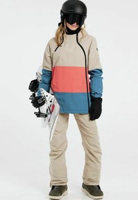 Snowboard-Ausrüstung mit einer beige-gestreiften Jacke in Blau und Koralle, beigen Hosen, schwarzen Handschuhen und einem schwarzen Helm, während die Snowboard-Bindungen in der Hand gehalten werden.