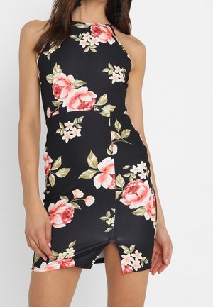 Frau trägt ein ärmelloses schwarzes Neckholderkleid mit großen rosa und weißen Blumenmustern und einem kleinen Schlitz vorne.