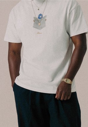Homme portant un T-shirt blanc avec un graphique d'une personne lisant un journal, pantalon foncé, montre en or et deux colliers en or sur fond uni.