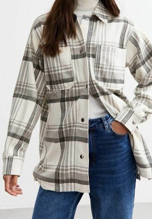 Woman wearing beige and gray plaid button-up shirt over white turtleneck, paired with blue jeans, posing with one hand in pocket.