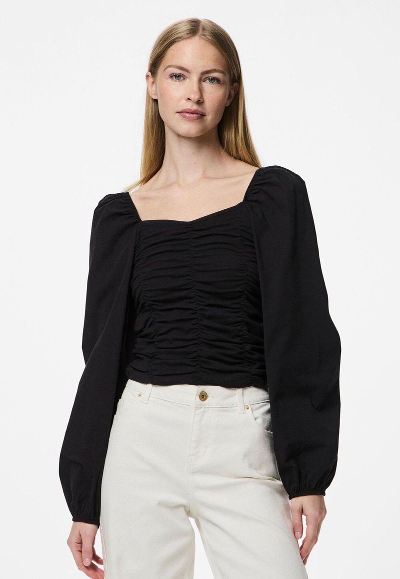 Blusa nera con scollatura quadrata, fronte arricciato e maniche lunghe a sbuffo; indossata con pantaloni avorio, con dettaglio di bottone in vita.