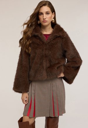 Bruine nepbont cropped jasje met een brede kraag en lange mouwen, gecombineerd met een geruite plissérok met rode accenten.