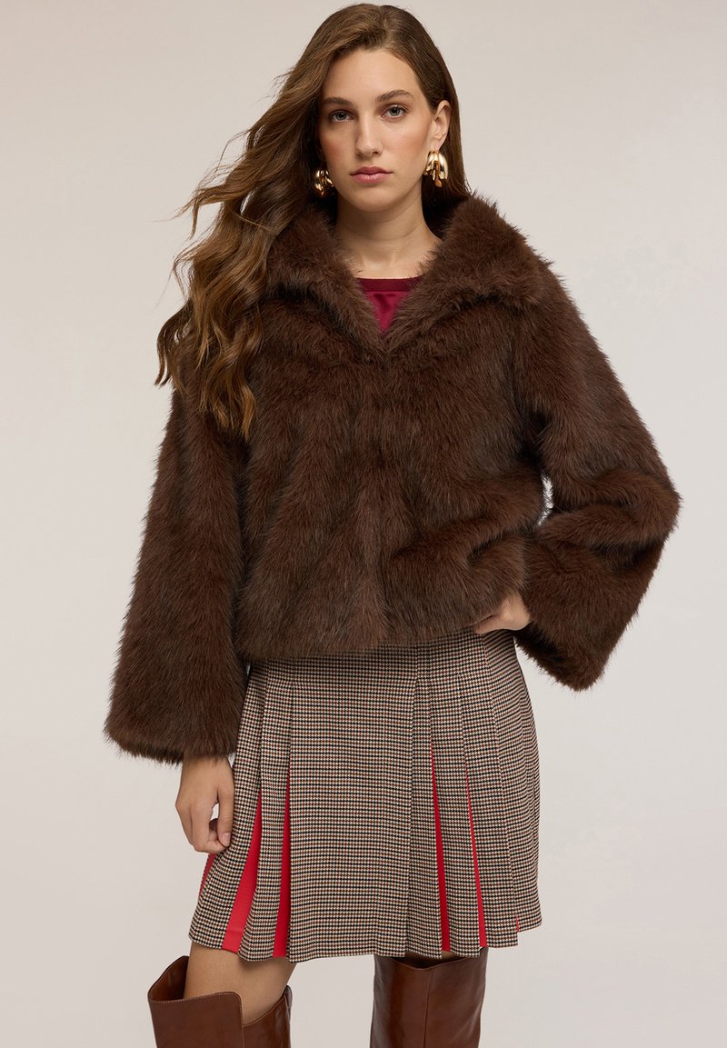 Giacca corta in finta pelliccia marrone con ampio colletto e maniche lunghe, abbinata a una gonna plissettata a scacchi con accenti rossi.