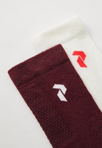 Par čarapa s rebrastim uzorkom, jedna maroon s bijelim logotipom, a druga bijela s crvenim logotipom, položene jedna preko druge na bijeloj površini.