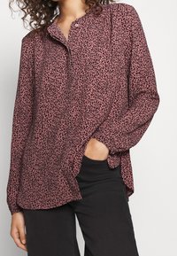 Blouse rose à manches longues avec imprimé léopard noir, design sans col, poche avant visible, poignets froncés et ourlet incurvé.