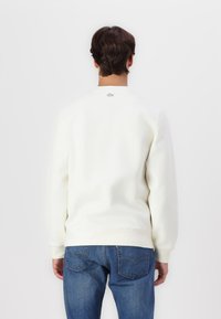 Sudadera blanca de algodón con cuello redondo y puños acanalados. Presenta un pequeño logo bordado en la parte posterior. Combinada con jeans de mezclilla azul.