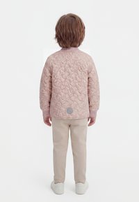 Otrok s kratkimi rjavimi lasmi, oblečen v roza prešito jakno, bež hlače in bele superge, stoji obrnjeno stran na beli podlagi.