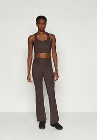 Kobieta w ciemno-brązowym bezrękawniku i dopasowanych spódniczkach w dzwon, w czarnych trampkach, stojąca na tle jednolitego jasnego tła.