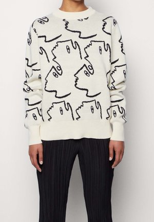 Pull blanc avec motif abstrait de visages en ligne continue noire, porté avec un pantalon plissé noir, montré du cou aux hanches.