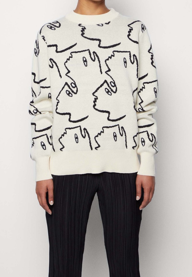 Pull blanc avec motif abstrait de visages en ligne continue noire, porté avec un pantalon plissé noir, montré du cou aux hanches.