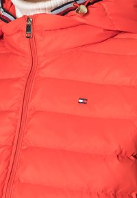 Nærbillede af en lys rød quiltet jakke med lynlås og et lille broderet firkantet logo i navy, hvidt og rødt på brystet.