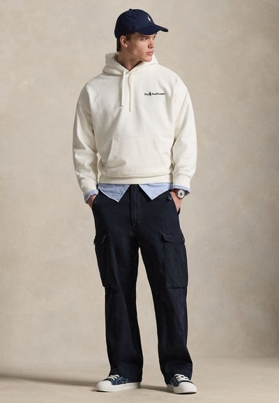 Λευκό βαμβακερό φούτερ με κουκούλα και μπροστινή τσέπη, σκούρο παντελόνι cargo, μπλε καπέλο και μπλε αθλητικά παπούτσια. Φορεμένο με ελαφρύ γαλάζιο πουκάμισο από κάτω.