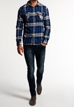 Chemise en flanelle à carreaux bleus et noirs avec deux poches poitrine, assortie à un jean denim foncé et des bottes grises, présentant une coupe ajustée.