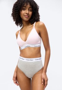 Γυναίκα με σγουρά μαλλιά φοράει ένα ανοιχτό ροζ σουτιέν Calvin Klein και γκρι εσώρουχο Calvin Klein με ψηλή μέση, στέκεται μπροστά σε απλό φόντο.