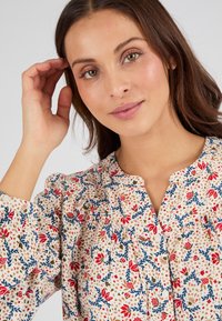 Blusa floreale con sfondo crema, caratterizzata da fiori rossi e foglie blu, scollatura rotonda, maniche a sbuffo e dettagli con bottoni.
