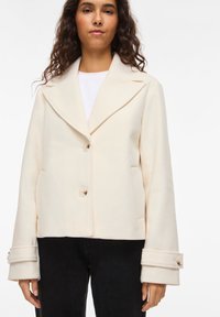 Εκρού cropped μπουφάν από απαλό ύφασμα, με γιακά με σχισμή, δύο κουμπιά και φαρδιά μανίκια με μανσέτες.